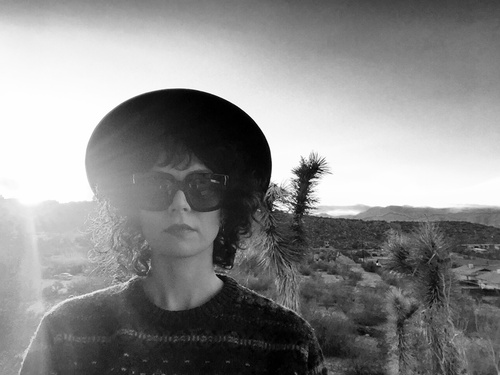 Sorenne in the desert at sunrise or sunset, wearing sunglasses and a wide-brimmed hat, Joshua trees and mountains behind her.